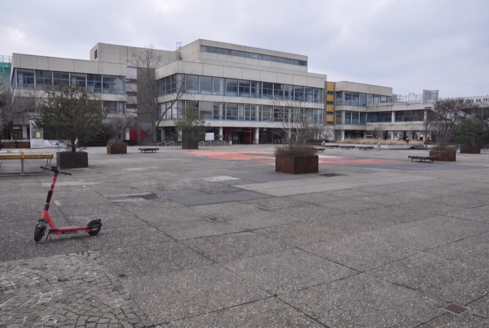KI-generierte Ansicht des Campusplatzes heute: grauer Belag, wenige Bäume, leere Bänke